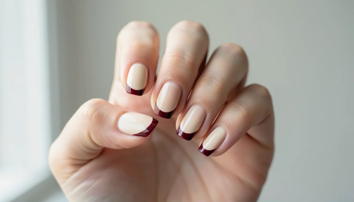 Mão feminina com unhas decoradas em estilo minimalista chic: esmalte bege cremoso com micro francesinha em vinho.